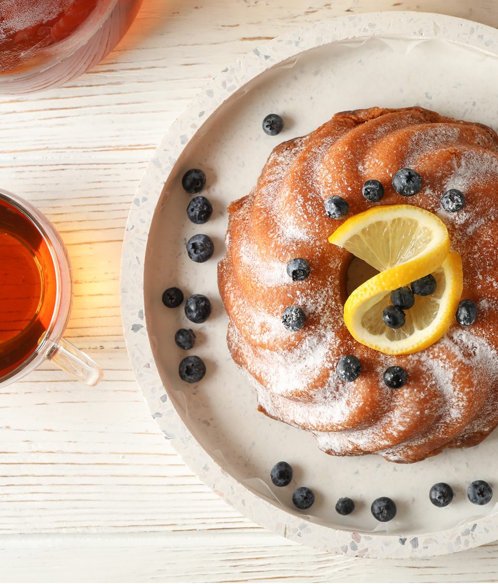 Gugelhopf mit Puderzucker, Heidelbeeren und Zitrone auf einem Teller neben einer Tasse Tee